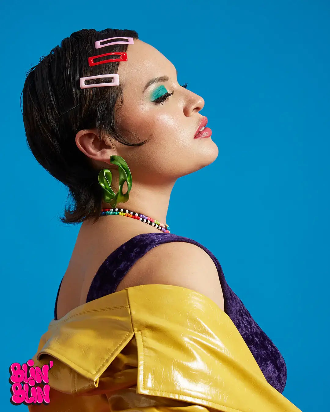 Mujer con aretes grandes verdes impresos en 3D, clips coloridos y chaqueta amarilla, fondo azul.